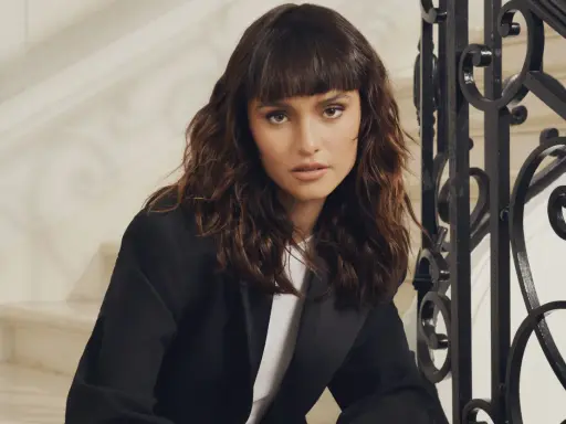 Une femme au regard intense arbore un **wolf cut cheveux bouclés** brun, porté avec un blazer noir.