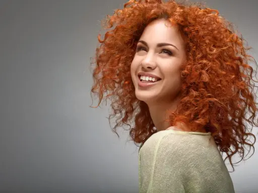 Une jeune femme au dégradé cheveux bouclés mi long, rousse, sourit joyeusement.