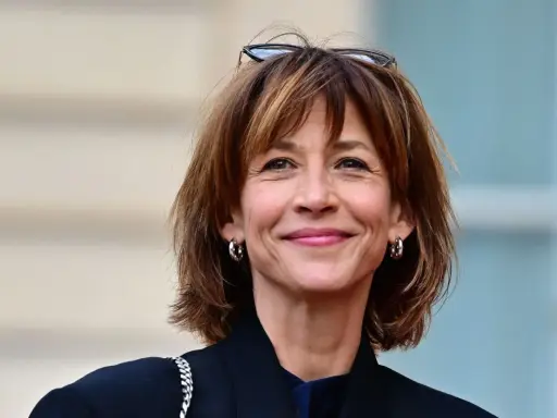 Une femme au sourire radieux arbore une coupe courte femme tendance, des lunettes sur la tête et des créoles argentées.