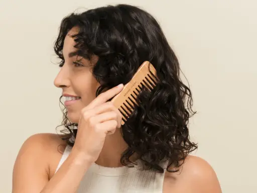 Une femme se peigne les cheveux bruns avec un peigne en bois. Elle sourit, car mes cheveux ne bouclent pas partout, mais ce peigne aide à définir les boucles.