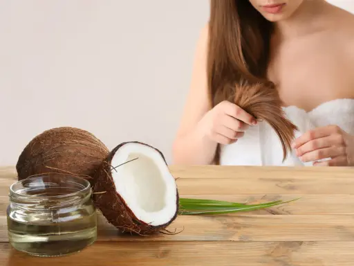 Un pot d'huile de coco et des noix de coco sur une table en bois, avec une femme en arrière-plan qui touche ses cheveux. L'huile de coco est idéale pour les cheveux bouclés.