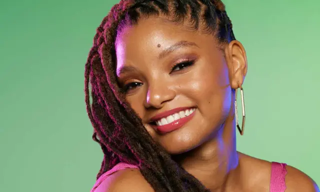 Jeune femme souriante avec une coiffure avec bijoux cheveux, des dreadlocks ornées de reflets violets, et de grandes boucles d'oreilles dorées.