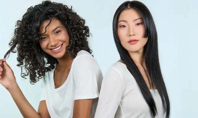 Deux femmes souriantes, l'une aux cheveux bouclés, l'autre aux cheveux lisses. La première joue avec ses mèches, évitant les cheveux fourchus jusqu'à la racine.