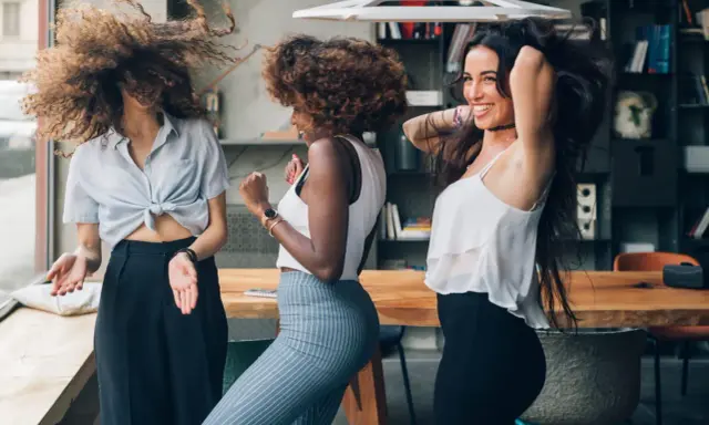 Trois femmes aux cheveux bouclés dansent joyeusement. Leurs chevelures volent, un exemple de comment avoir de belles boucles sans frisottis.