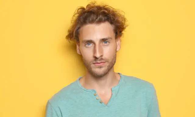 Un jeune homme aux cheveux bouclés et aux yeux bleus regarde la caméra sur fond jaune.
