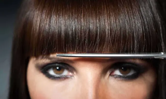 Un modèle de coiffure aux yeux marron et au maquillage prononcé, une frange brune est coupée par des ciseaux.