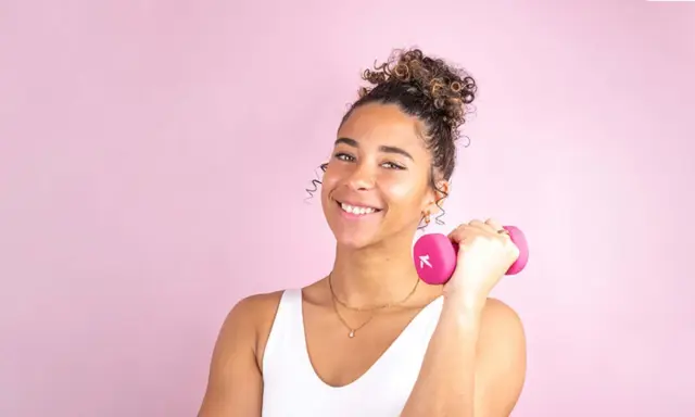 Une jeune femme souriante avec une coiffure sport cheveux bouclés tient une haltère rose.
