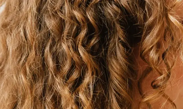 Une femme se coiffe les cheveux ondulés le matin, révélant des boucles dorées et un sourire radieux.