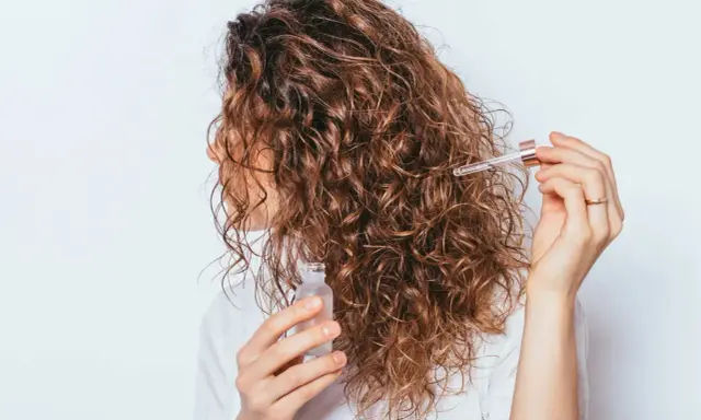 Une femme applique une huile capillaire sur ses cheveux frisottis sur le dessus. Ses boucles brunes brillent sous la lumière.