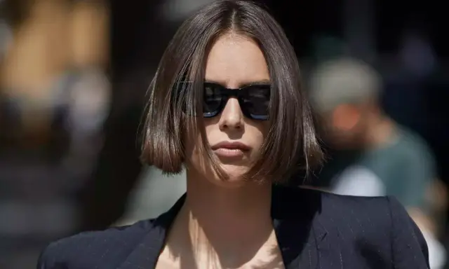 Une femme au regard intense arbore une coupe carré droit dégradé, accentuée par des lunettes de soleil noires et un blazer chic.