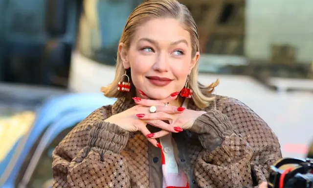 Gigi Hadid, avec ses boucles d'oreilles en forme de dé rouge, pose avec un sourire malicieux. Son carré plaqué en arrière et son manteau texturé complètent son look.