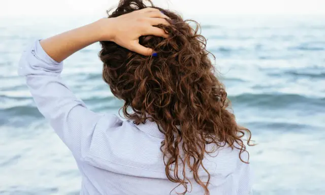 Main tenant ses cheveux bouclés, une femme en chemise rayée profite de la brise marine. Elle n'a pas peur de brosser ses cheveux bouclés tous les jours.