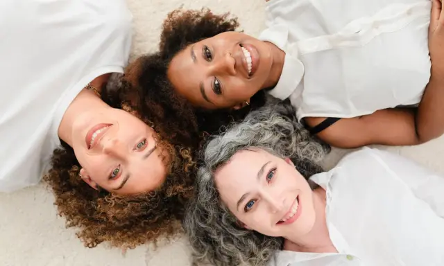 Trois femmes souriantes, aux cheveux bouclés, frisés et gris, partagent un moment complice. Un scruncher cheveux pourrait parfaire leur look.