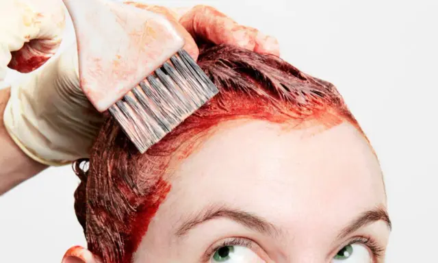Application de teinture rouge sur les cheveux, étape clé de la routine cheveux colorés. Une main gantée utilise un pinceau pour une application précise.