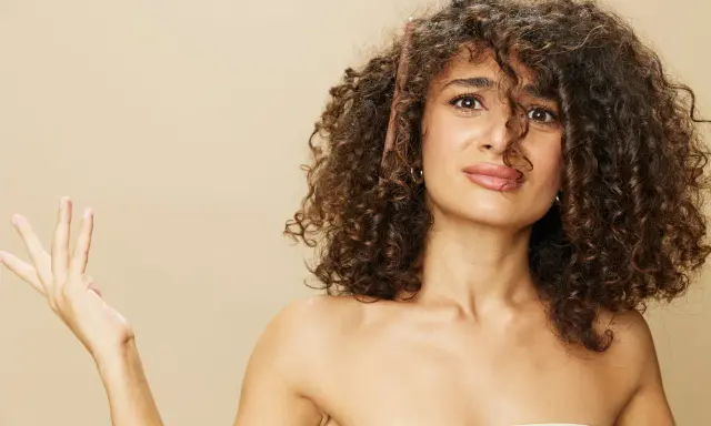 Une femme aux cheveux longs et bouclés, une brosse à cheveux coincée dedans, lève une main ouverte.