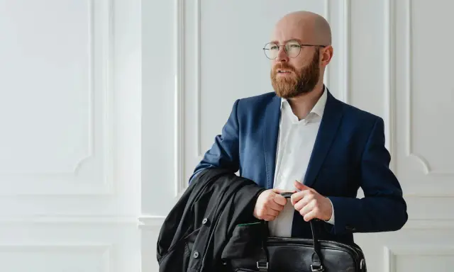 Un homme crane rasé, barbe rousse, lunettes, en costume bleu, porte une veste noire et une mallette.