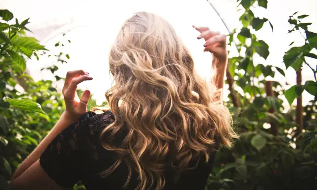 Chevelure blonde ondulée, baignée de lumière, entourée de verdure. Une scène évoquant la douceur d'une **décoloration végétale** naturelle.