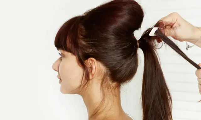 Une main ajuste une mèche pour créer un chignon sophistiqué. La coiffure volumineuse est en cours de réalisation.