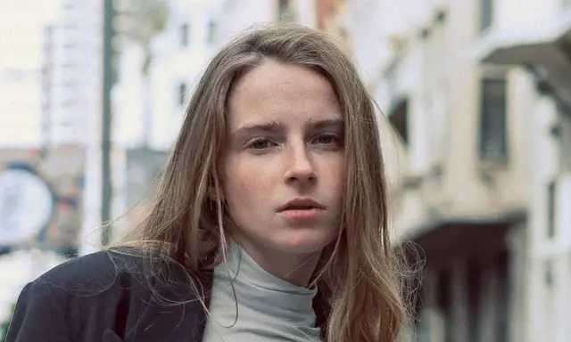 Jeune femme au regard intense, ses cheveux châtain clair cendré ou glacé encadrent son visage parsemé de taches de rousseur. Elle porte un col roulé gris et une veste sombre.