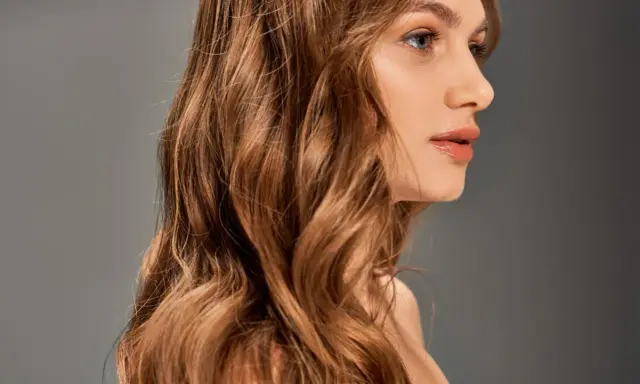 Chevelure ondulée, passant du blond au chatain, d'une femme au profil délicat, sur fond gris.