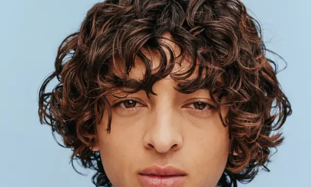 Un jeune homme au regard intense arbore une magnifique coupe cheveux bouclés homme, des boucles brunes et rebelles encadrant son visage.