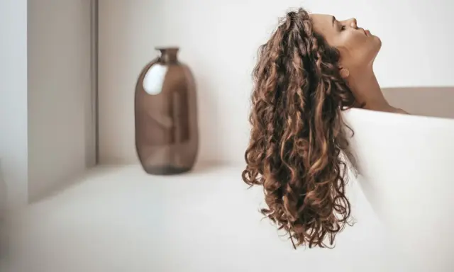 Une femme aux cheveux bouclés se détend dans une baignoire. Elle se demande combien de shampoing par lavage est idéal pour ses cheveux.