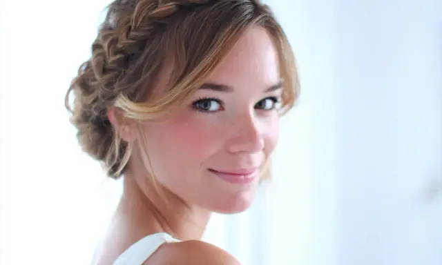 Une jeune femme au sourire doux arbore une coiffure demi tresse élégante, ses cheveux blonds ornés d'une tresse subtile.