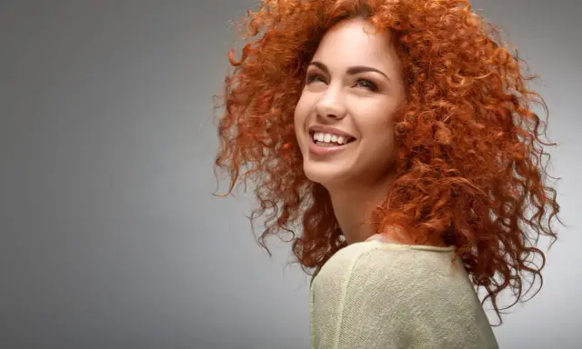 Une jeune femme au dégradé cheveux bouclés mi long, rousse, sourit joyeusement.