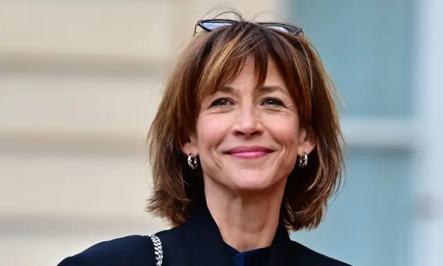 Une femme au sourire radieux arbore une coupe courte femme tendance, des lunettes sur la tête et des créoles argentées.