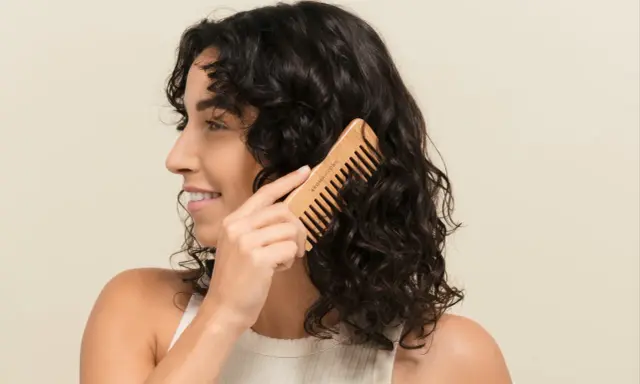Une femme se peigne les cheveux bruns avec un peigne en bois. Elle sourit, car mes cheveux ne bouclent pas partout, mais ce peigne aide à définir les boucles.