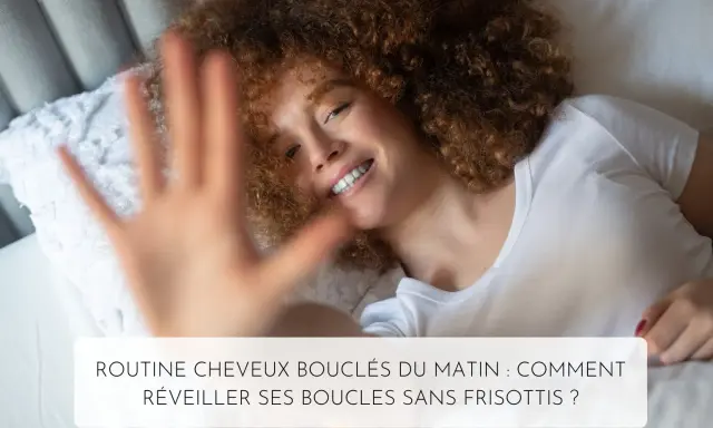 Une femme souriante aux cheveux 3c bouclés se réveille dans son lit. Sa main est devant l'objectif.