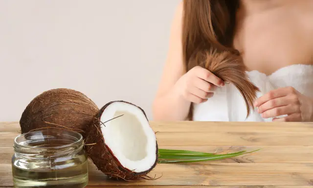 Un pot d'huile de coco et des noix de coco sur une table en bois, avec une femme en arrière-plan qui touche ses cheveux. L'huile de coco est idéale pour les cheveux bouclés.