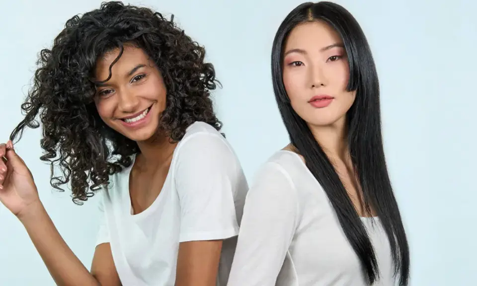 Deux femmes souriantes, l'une aux cheveux bouclés, l'autre aux cheveux lisses. La première joue avec ses mèches, évitant les cheveux fourchus jusqu'à la racine.