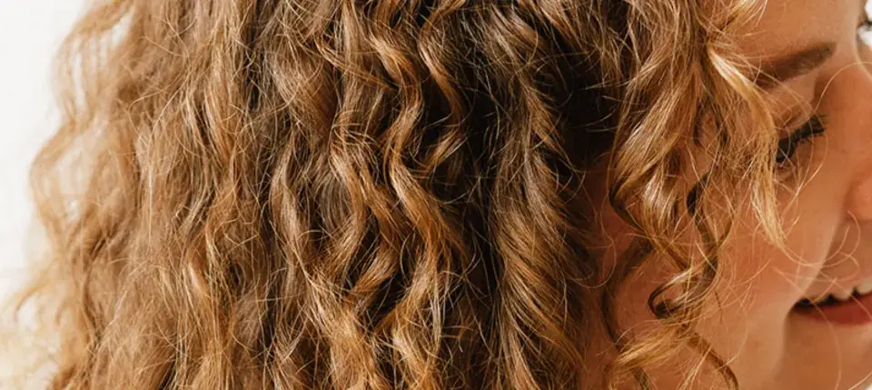 Une femme se coiffe les cheveux ondulés le matin, révélant des boucles dorées et un sourire radieux.
