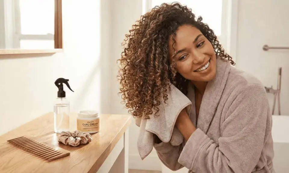 Une femme souriante utilise une serviette sur ses cheveux bouclés. Sur la table, un peigne, une crème pour boucles et un spray d'eau pour un tuto coiffure cheveux bouclés naturel.