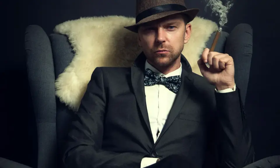 Homme au costume élégant, chapeau et nœud papillon, avec une coupe cheveux rockabilly homme. Il fume un cigare, assis dans un fauteuil.