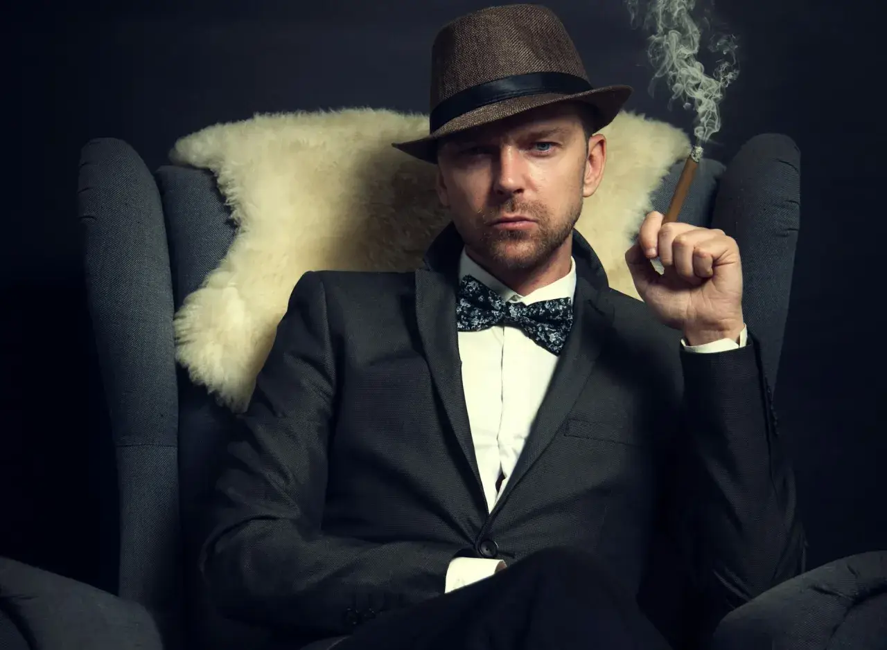 Homme au costume élégant, chapeau et nœud papillon, avec une coupe cheveux rockabilly homme. Il fume un cigare, assis dans un fauteuil.