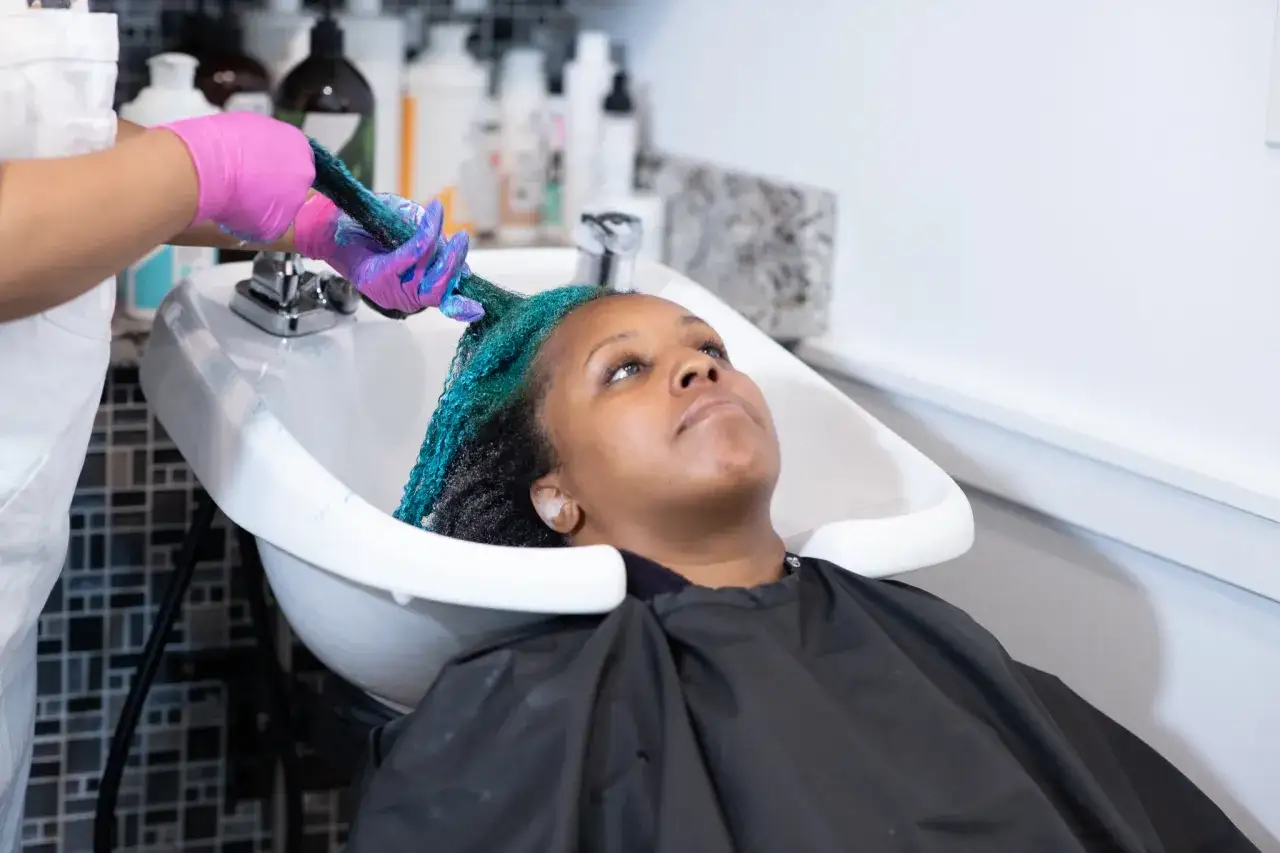 Une personne en gants roses applique un soin bleu turquoise sur les cheveux d'une femme dans une bac de lavage. C'est une étape clé pour savoir comment bien rincer ses cheveux.