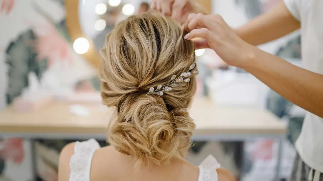Une mariée se fait coiffer un chignon bas orné d'une barrette scintillante. Ses cheveux blonds sont tressés avec élégance.