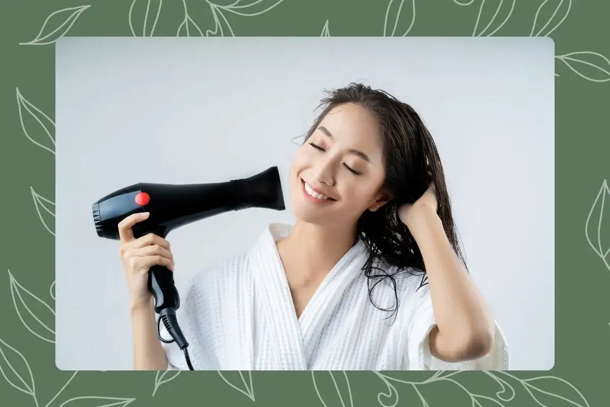 Une femme souriante sèche ses cheveux humides avec un sèche-cheveux, vêtue d'un peignoir blanc.