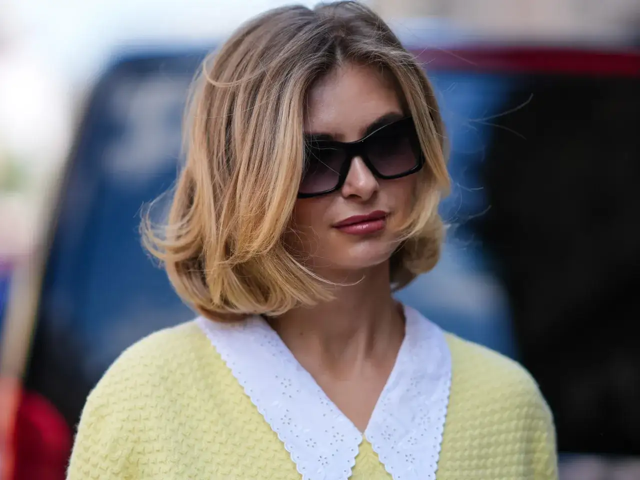 Une femme arbore une coupe carré droit dégradé, des lunettes de soleil oversize et un pull jaune à col blanc brodé.