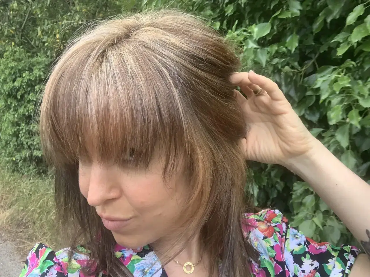 Cheveux ch&acirc;tains avec des reflets blonds, r&eacute;sultat d'un &eacute;claircir cheveux naturellement. Une femme touche ses cheveux devant un mur de verdure.