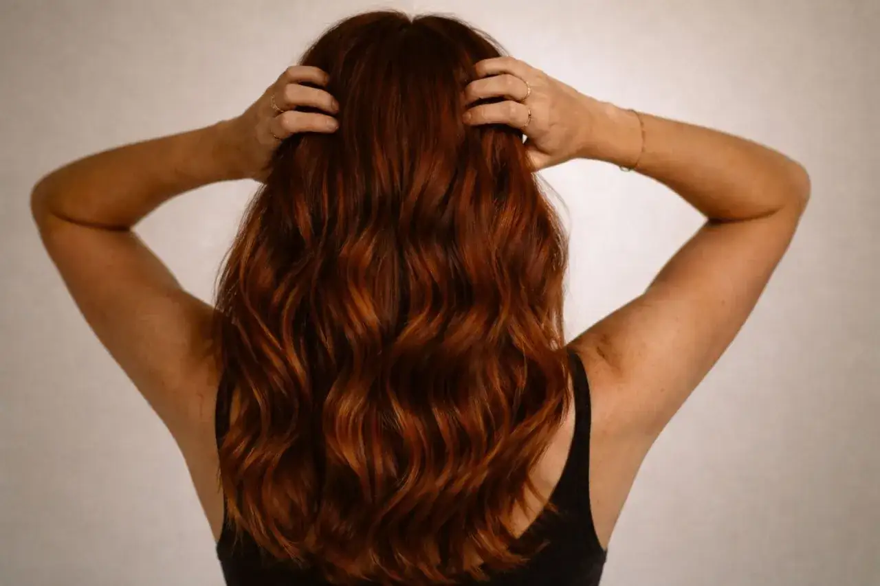 Vue arri&egrave;re d'une femme aux cheveux bruns ondul&eacute;s, touchant sa chevelure avec ses mains orn&eacute;es de bagues.