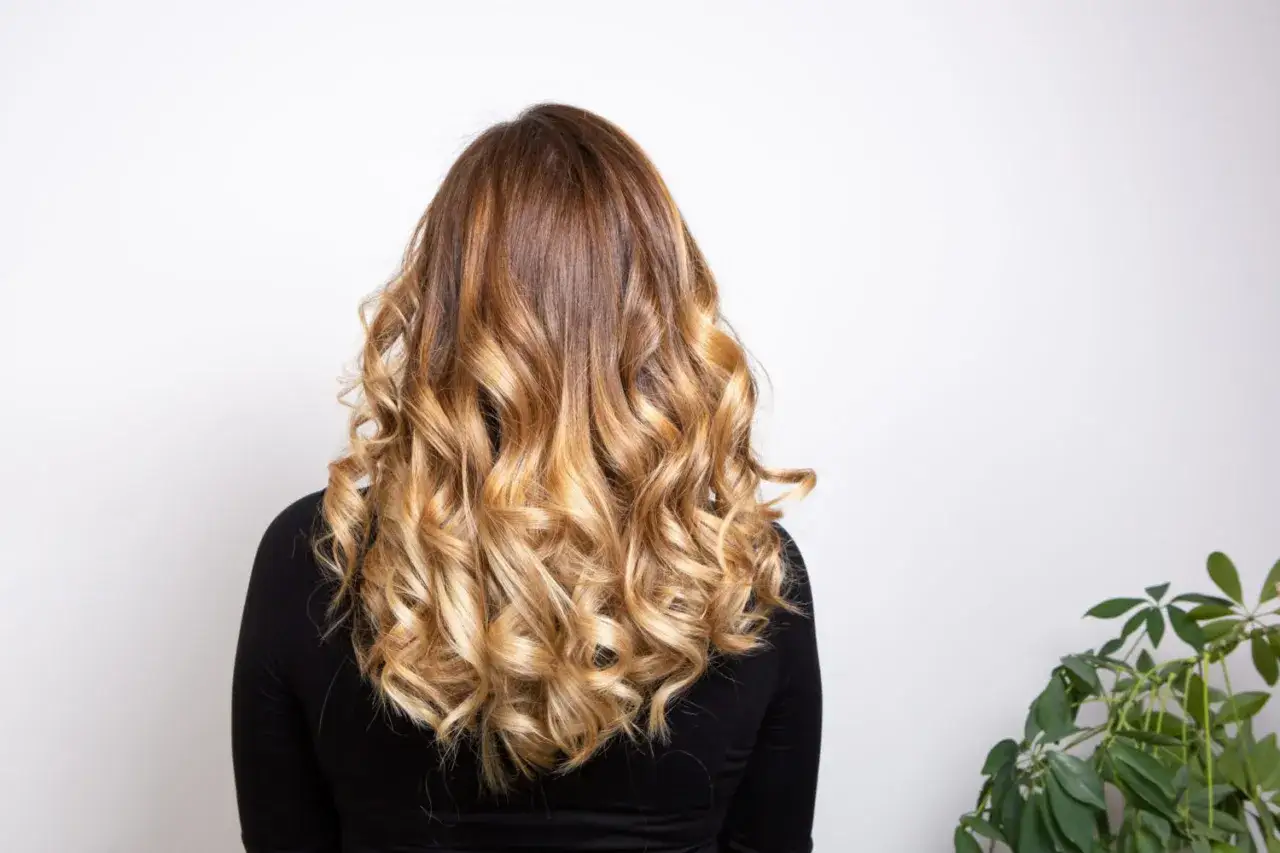 Chevelure ondul&eacute;e avec un balayage chatain clair subtil, cr&eacute;ant un effet ensoleill&eacute; naturel.