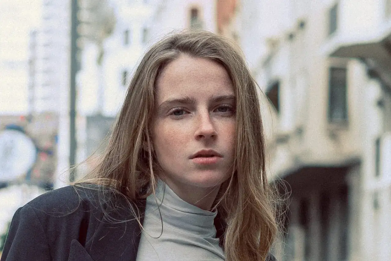 Jeune femme au regard intense, ses cheveux châtain clair cendré ou glacé encadrent un visage parsemé de taches de rousseur. Elle porte un col roulé gris et une veste sombre.
