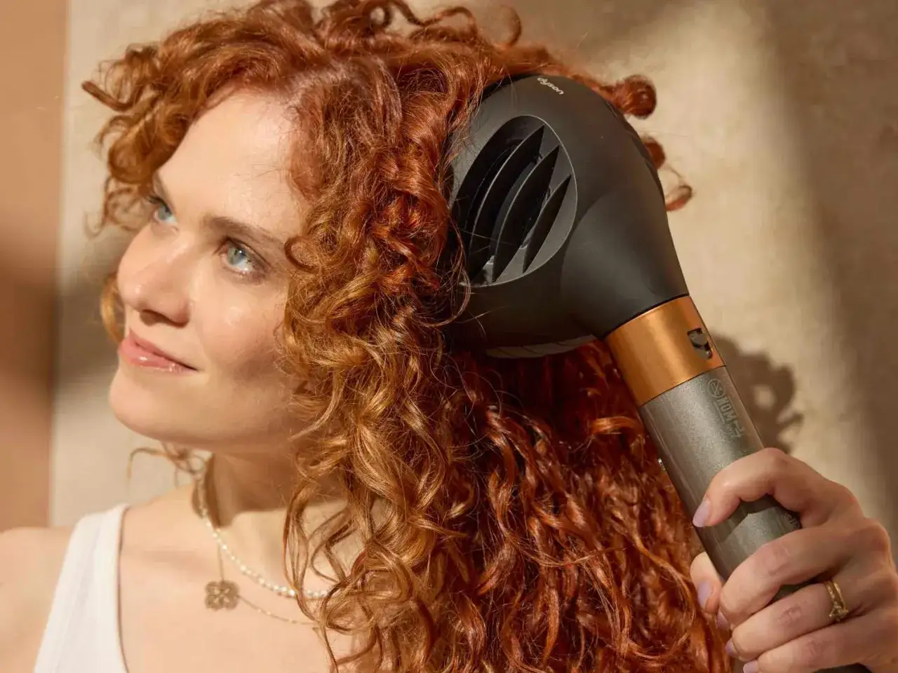Femme aux cheveux roux et bouclés utilisant un sèche-cheveux Dyson pour coiffer ses cheveux frisottis sur le dessus.