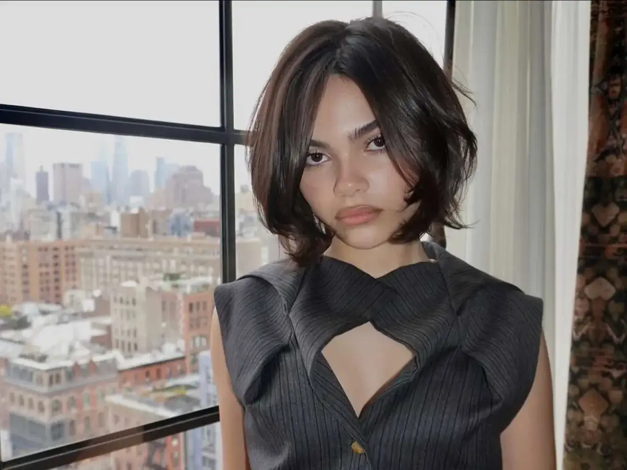 Jeune femme avec un carré plongeant court, ses cheveux fins encadrent son visage. Elle porte une tenue grise stylée devant une fenêtre avec vue sur la ville.