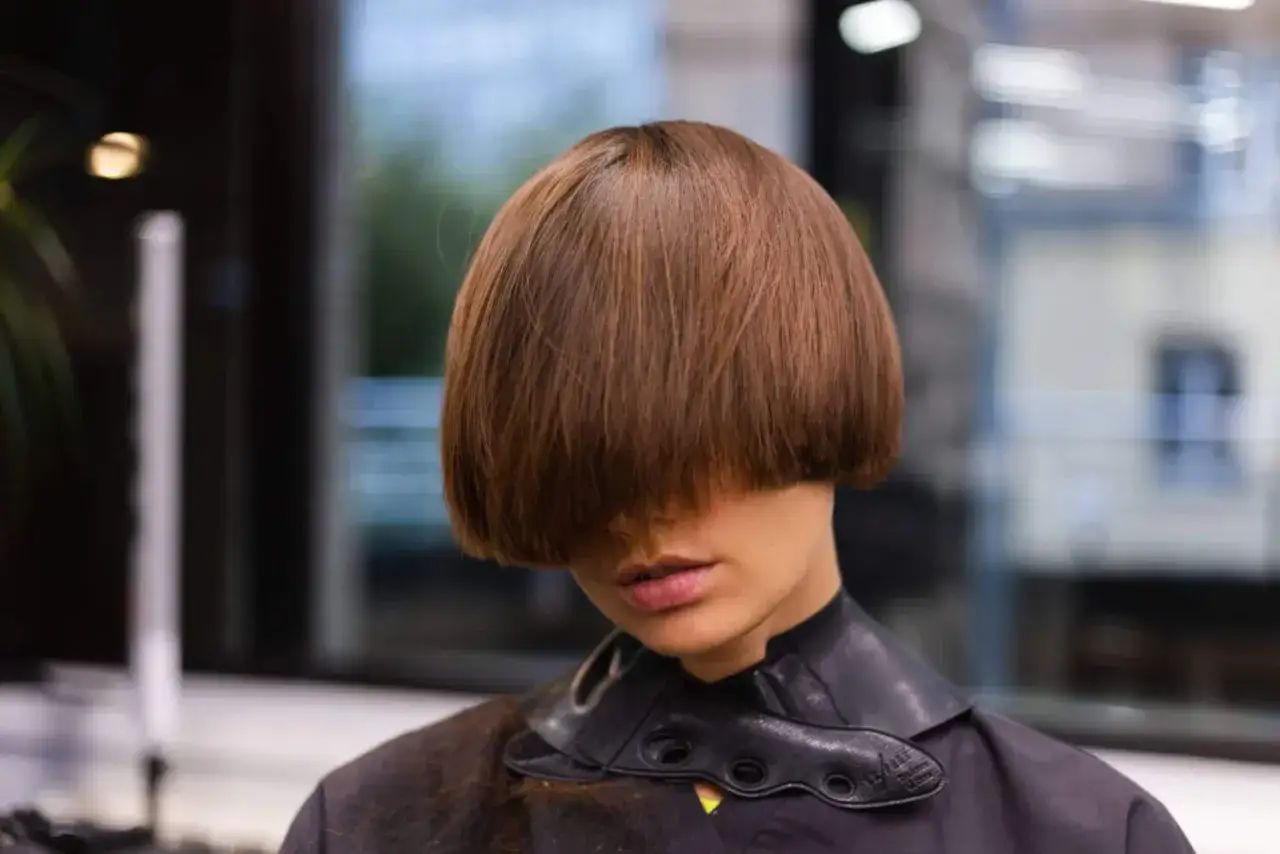 Une femme arbore une coupe au bol audacieuse, ses cheveux ch&acirc;tains cachant son regard. Le col noir de sa tenue ajoute une touche moderne.