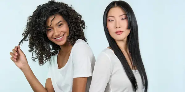 Deux femmes souriantes, l'une aux cheveux bouclés, l'autre aux cheveux lisses. La première joue avec ses mèches, évitant les cheveux fourchus jusqu'à la racine.