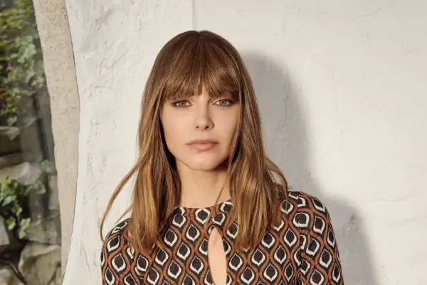 Une femme au carré frange rideau brune, aux yeux verts perçants, porte une robe à motifs géométriques.